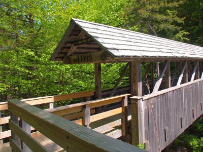 [Image taken on the trail just before the all wooden covered bridge showing the date and name of the bridge on the roof portion under which people walk.]